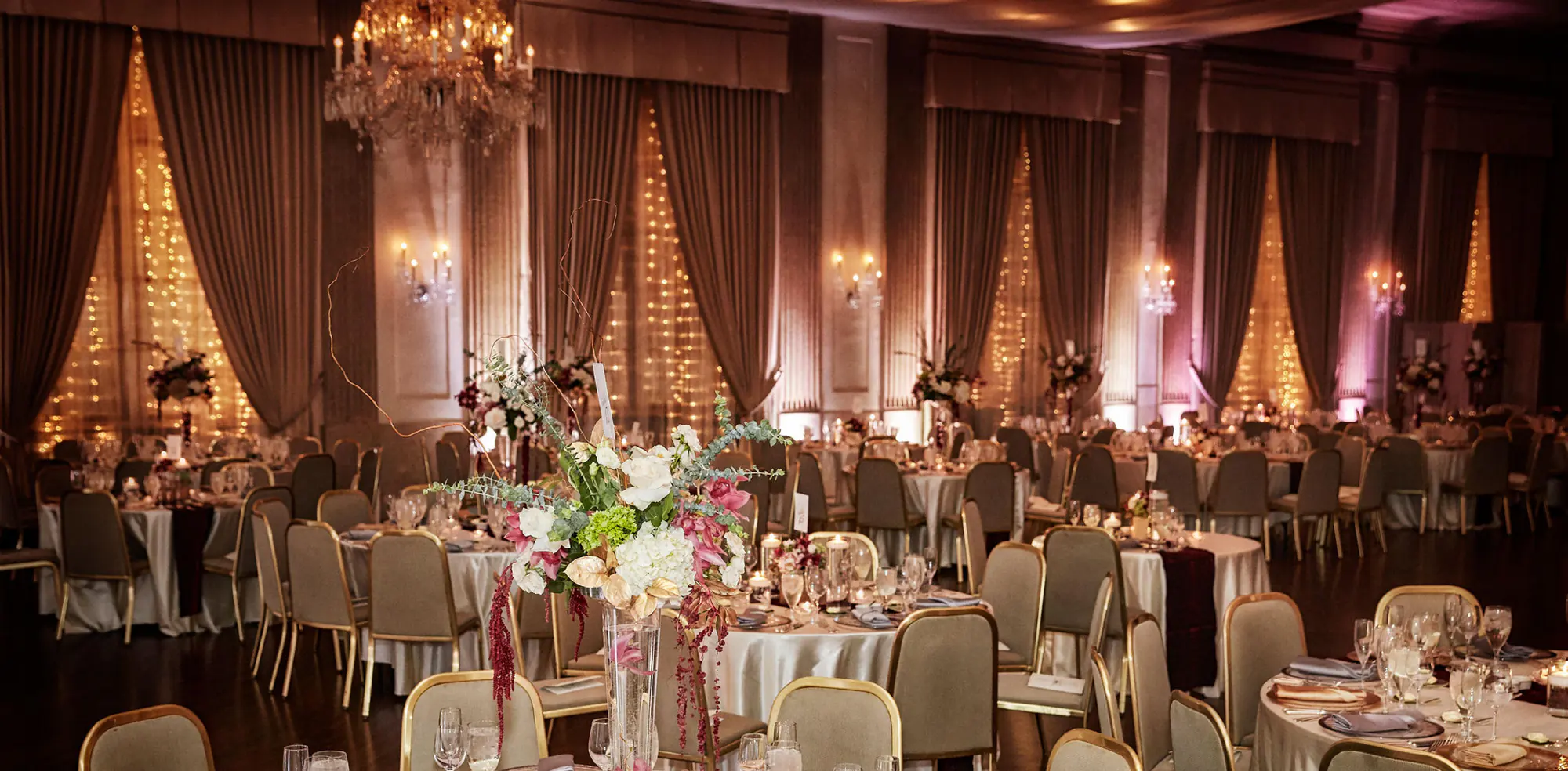 A Standard Club ballroom in the evening, round tables and gilt chairs beneath a crystal chandelier, tall floral centrepieces, and curtained windows hung with strings of small lights.