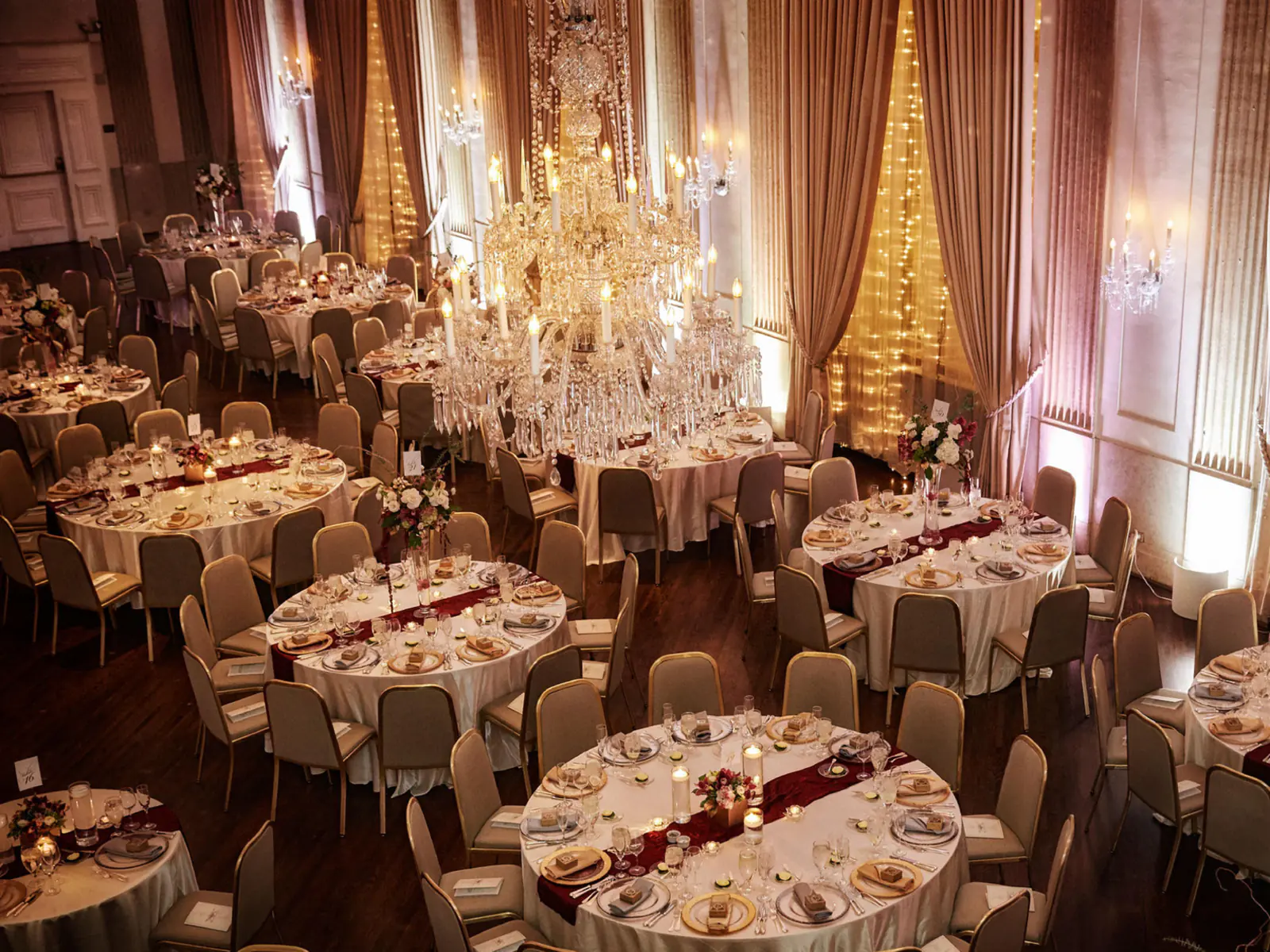 A Standard Club ballroom seen from above, set for a dinner: round tables with candles and flowers beneath a crystal chandelier, curtains lit with strings of small lights.