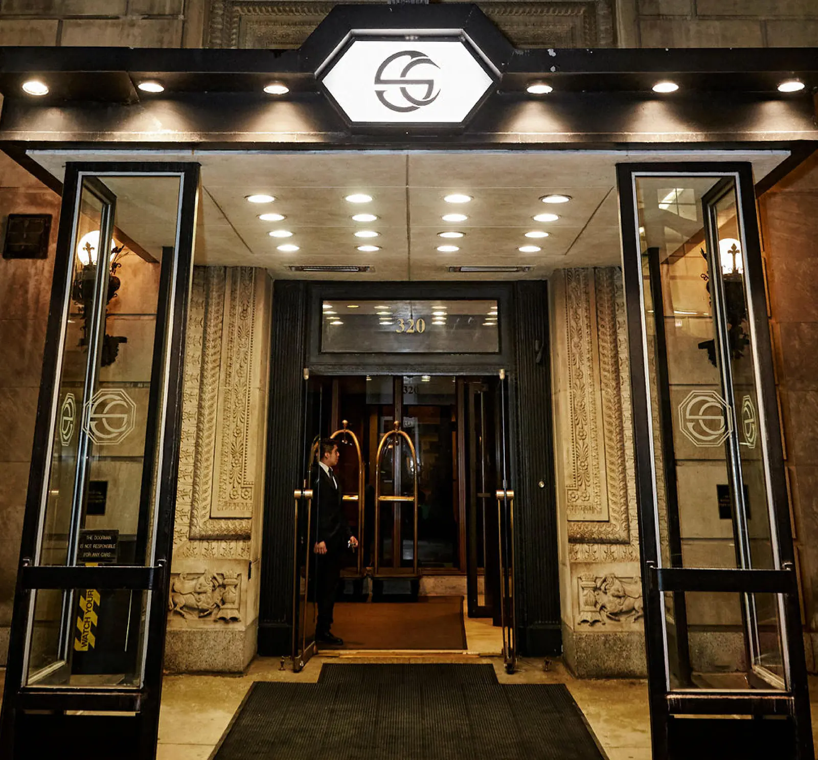 The entrance of the Standard Club at 320 South Plymouth Court at night, its lit canopy bearing the Club’s octagonal logo above open glass doors set in a carved stone facade.