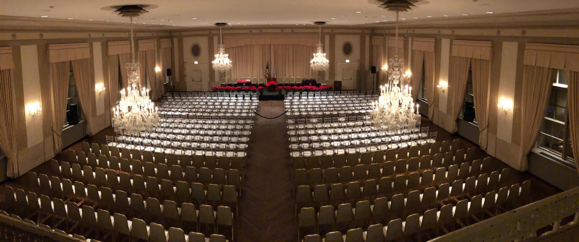 The Standard Club’s Grand Ballroom from the balcony, rows of empty chairs divided by a centre aisle leading to a stage, crystal chandeliers hanging above.