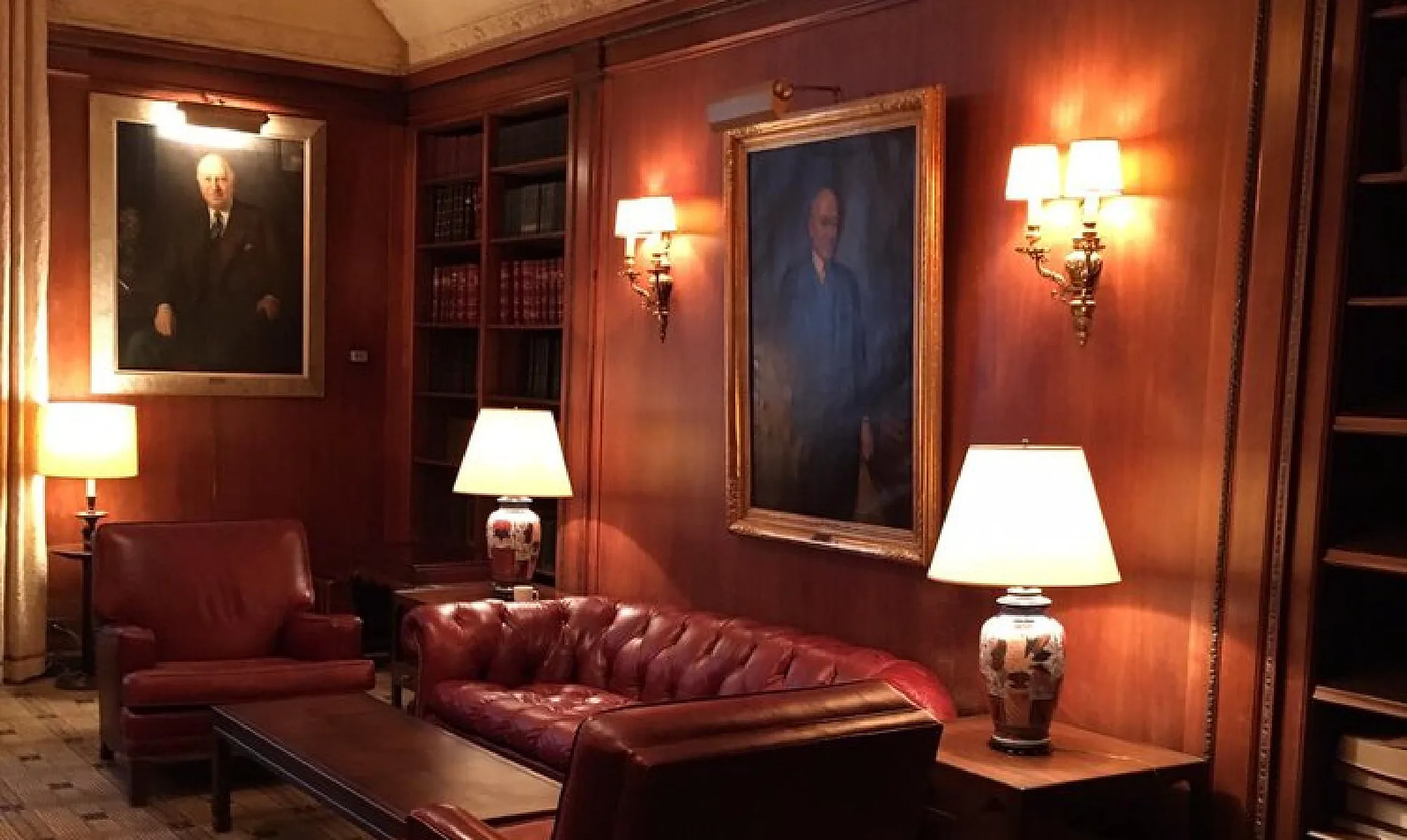 The Standard Club library: a red leather sofa and armchair beside a low table, two table lamps and a pair of brass wall sconces lighting wood panelling, a bookcase of bound volumes, and two framed portraits of men in suits.